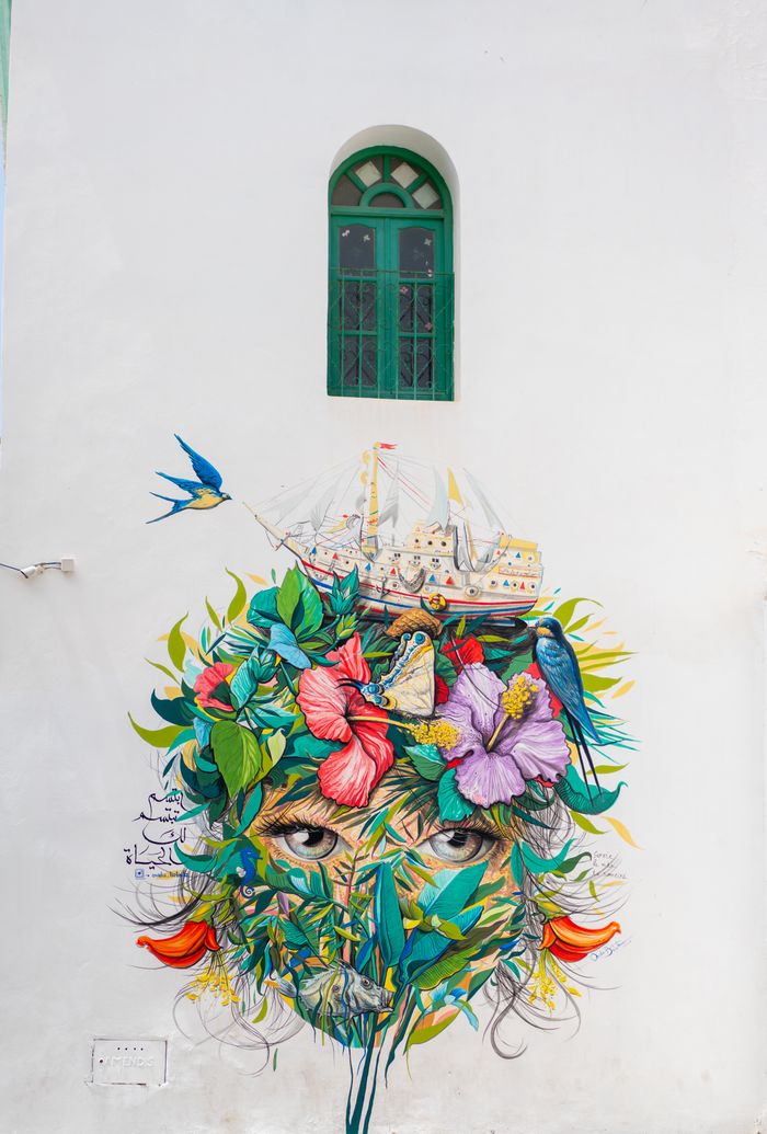 About Us Colorful mural with floral elements and birds on a white wall under a green window.